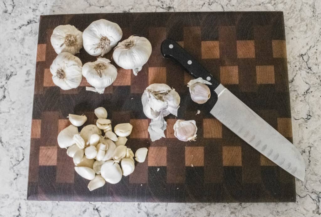 A beautiful wood cutting board with a knife and some fresh garlic in the process of being peeled. 