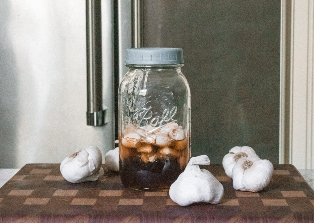 A glass jar containing honey and garlic in the process of fermenting, surrounded by fresh garlic heads. 